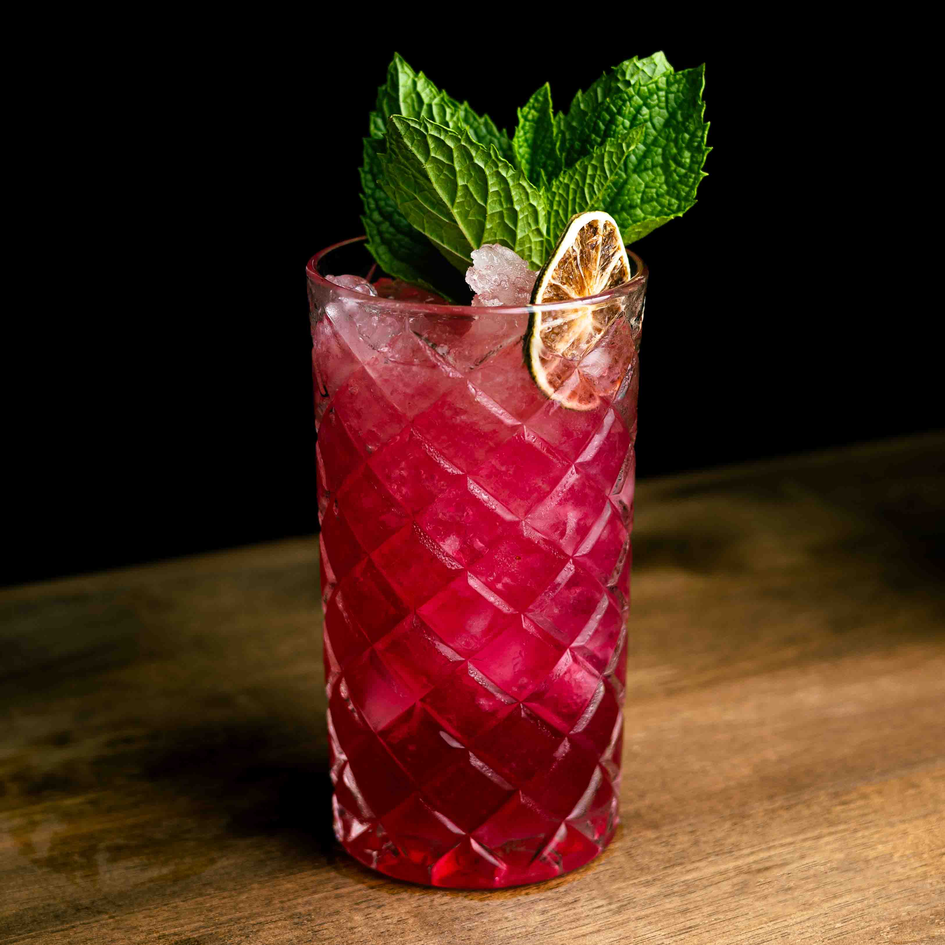 Red cocktail with lime and mint in a textured glass on a wooden surface
