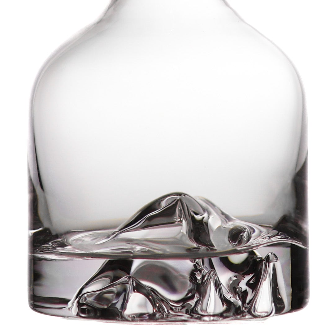 Clear glass decanter with a sculptural mountain-like base, shown close up on a white background.