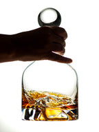 Hand holding a glass decanter with whiskey and ice cubes on a white background