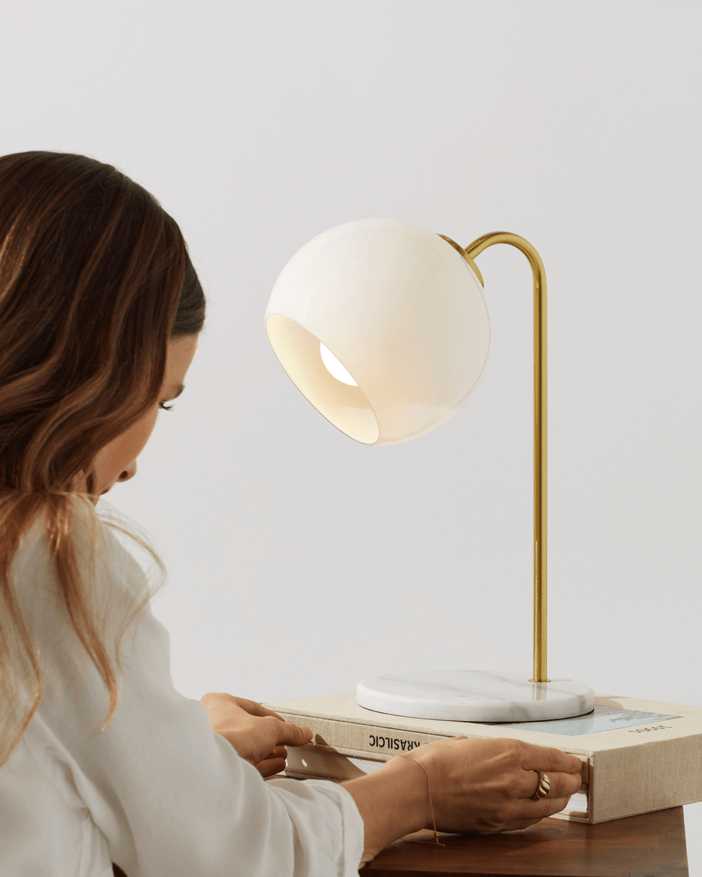 Woman reading a book under a modern desk lamp on a wooden surface.