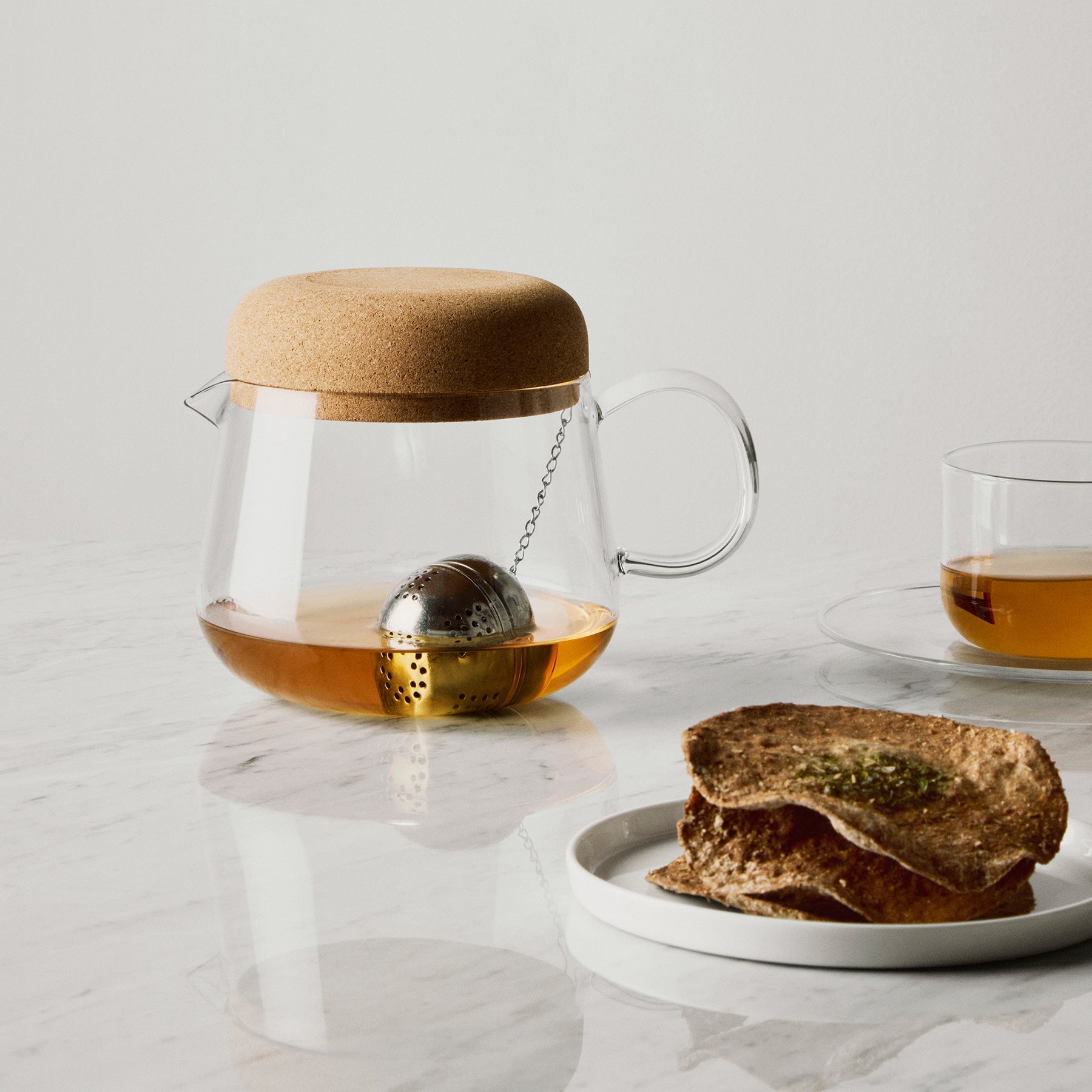 Clear glass teapot with cork lid, tea ball, and glass of tea on a marble surface.