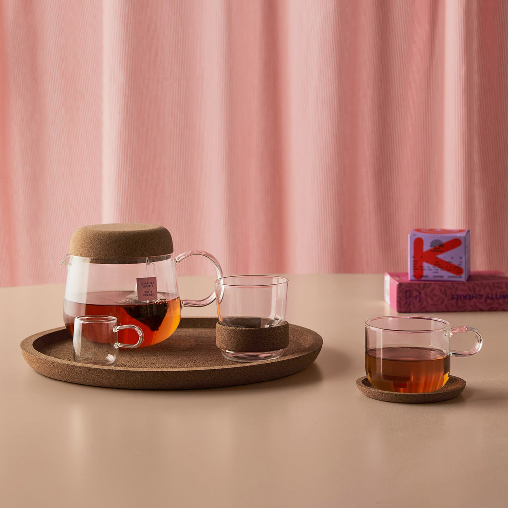 Tea-making set with glass teapot, cups, and a wooden tray on a pink curtain background