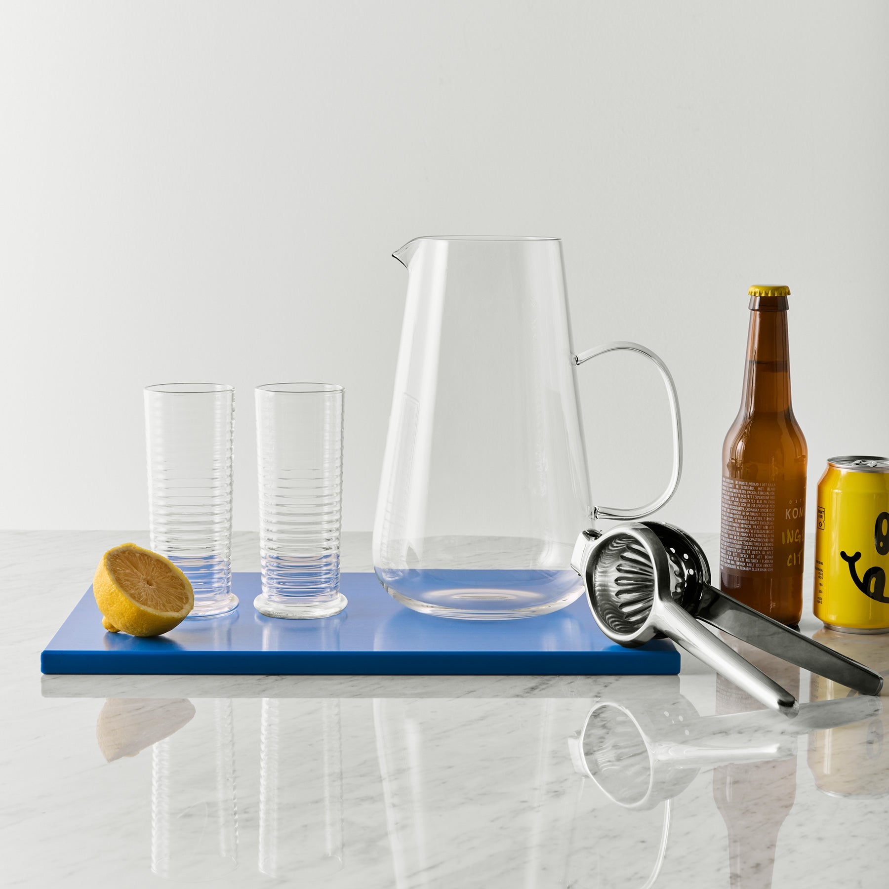 Clear pitcher, glasses, lemon, beer bottle, and can on a blue cutting board with a white background
