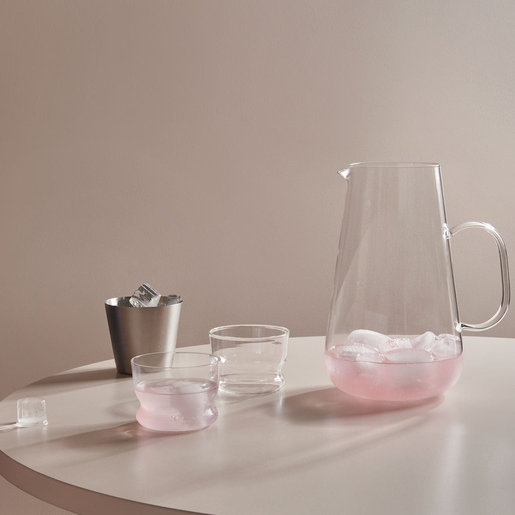 Clear glass pitcher and cups with pink liquid on a white surface against a beige wall.