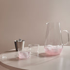 Clear glass pitcher and cups with pink liquid on a white surface against a beige wall.