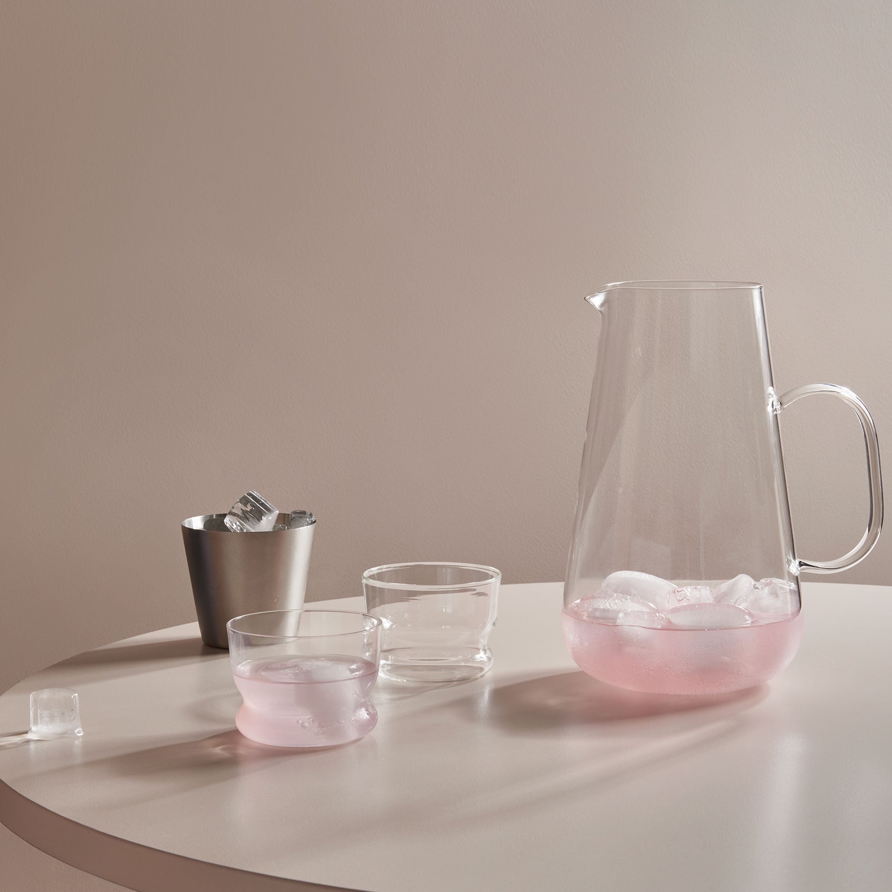 Clear glass pitcher and cups with pink liquid on a white surface against a beige wall.