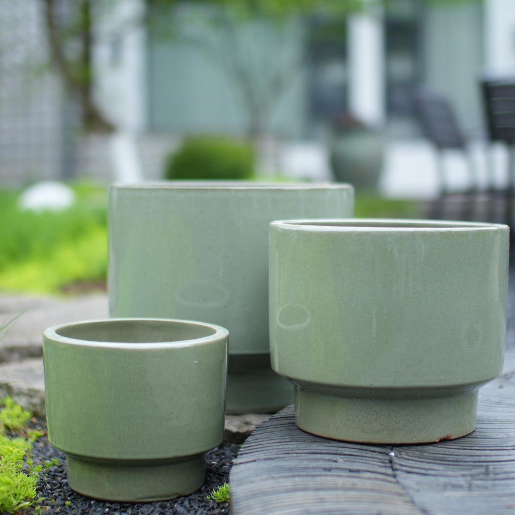 Three light sage green ceramic planters in different sizes arranged on a weathered wooden surface outdoors, with a blurred garden background.