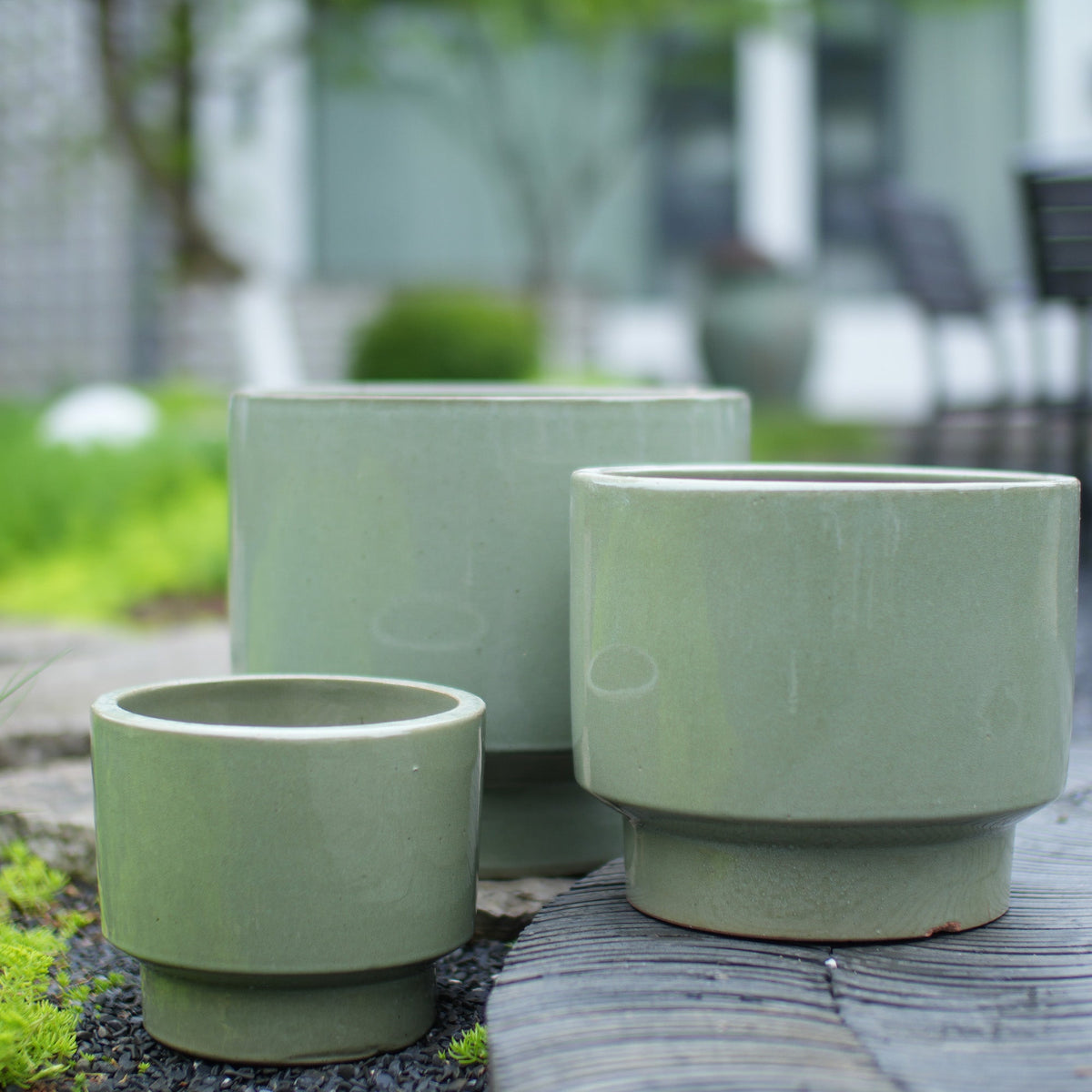 Three light sage green ceramic planters in different sizes arranged on a weathered wooden surface outdoors, with a blurred garden background.