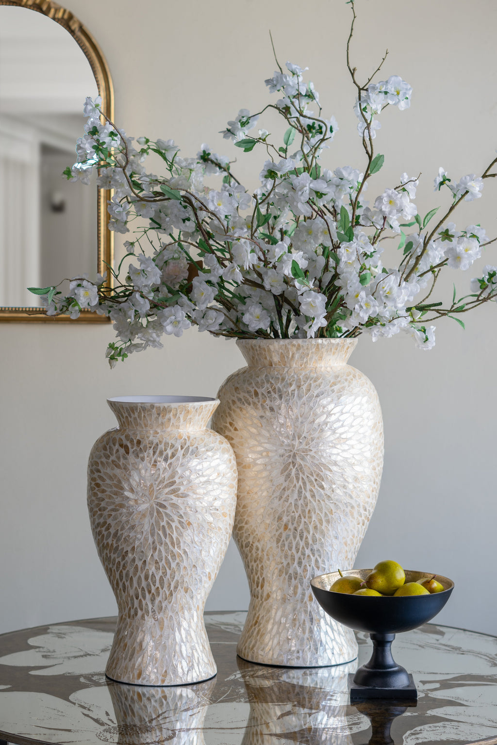 Two textured cream ceramic vases filled with white blossoms and greenery, displayed on a reflective table next to a black pedestal bowl of olives in an elegant interior setting.