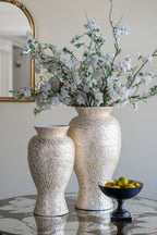Two textured cream ceramic vases filled with white blossoms and greenery, displayed on a reflective table next to a black pedestal bowl of olives in an elegant interior setting.