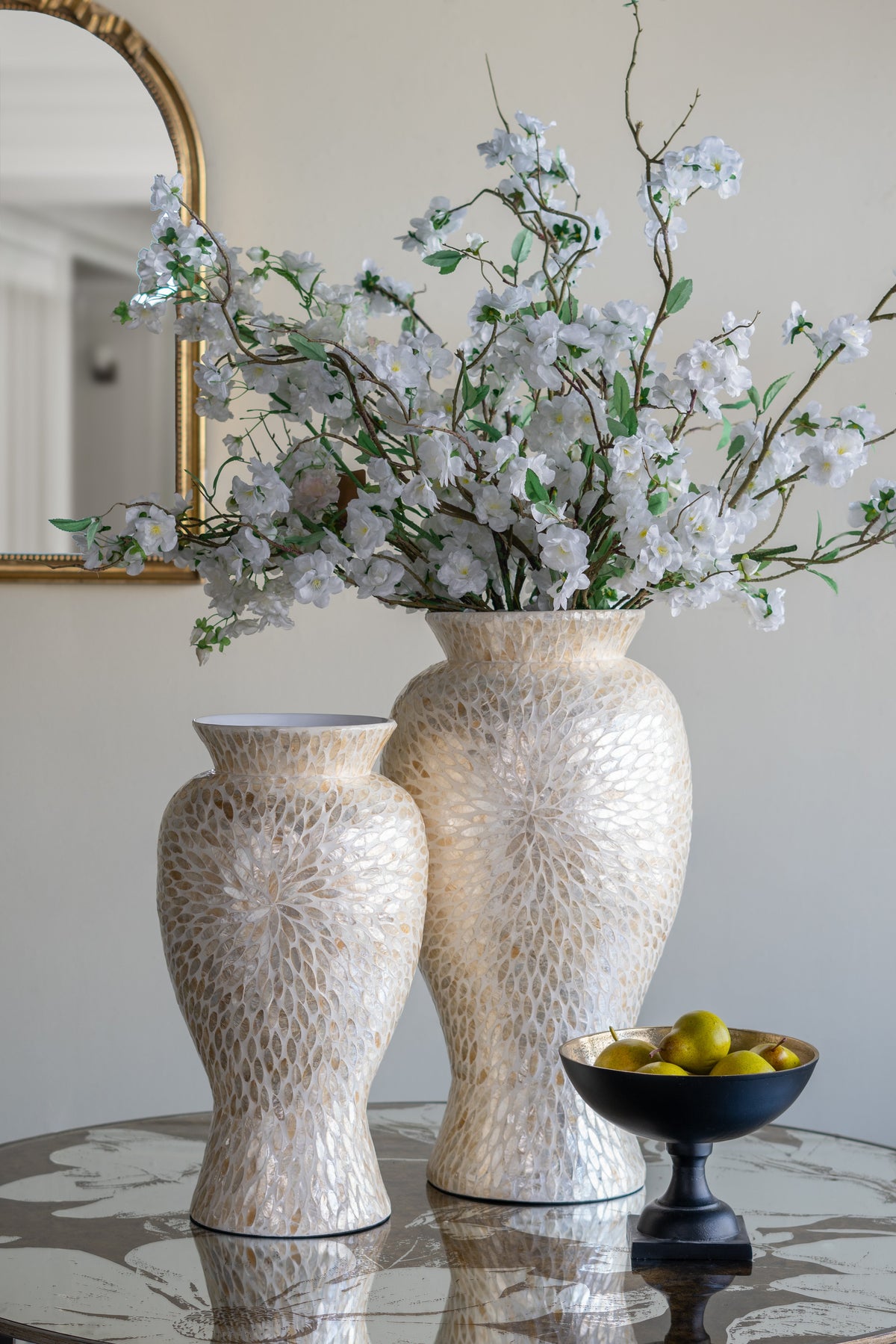 Two textured cream ceramic vases filled with white blossoms and greenery, displayed on a reflective table next to a black pedestal bowl of olives in an elegant interior setting.