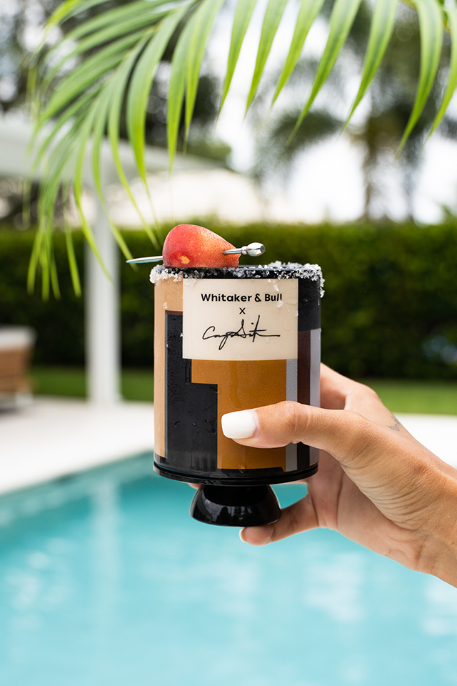 Hand holding a cocktail with a 'Whitaker & Bull' label by a poolside
