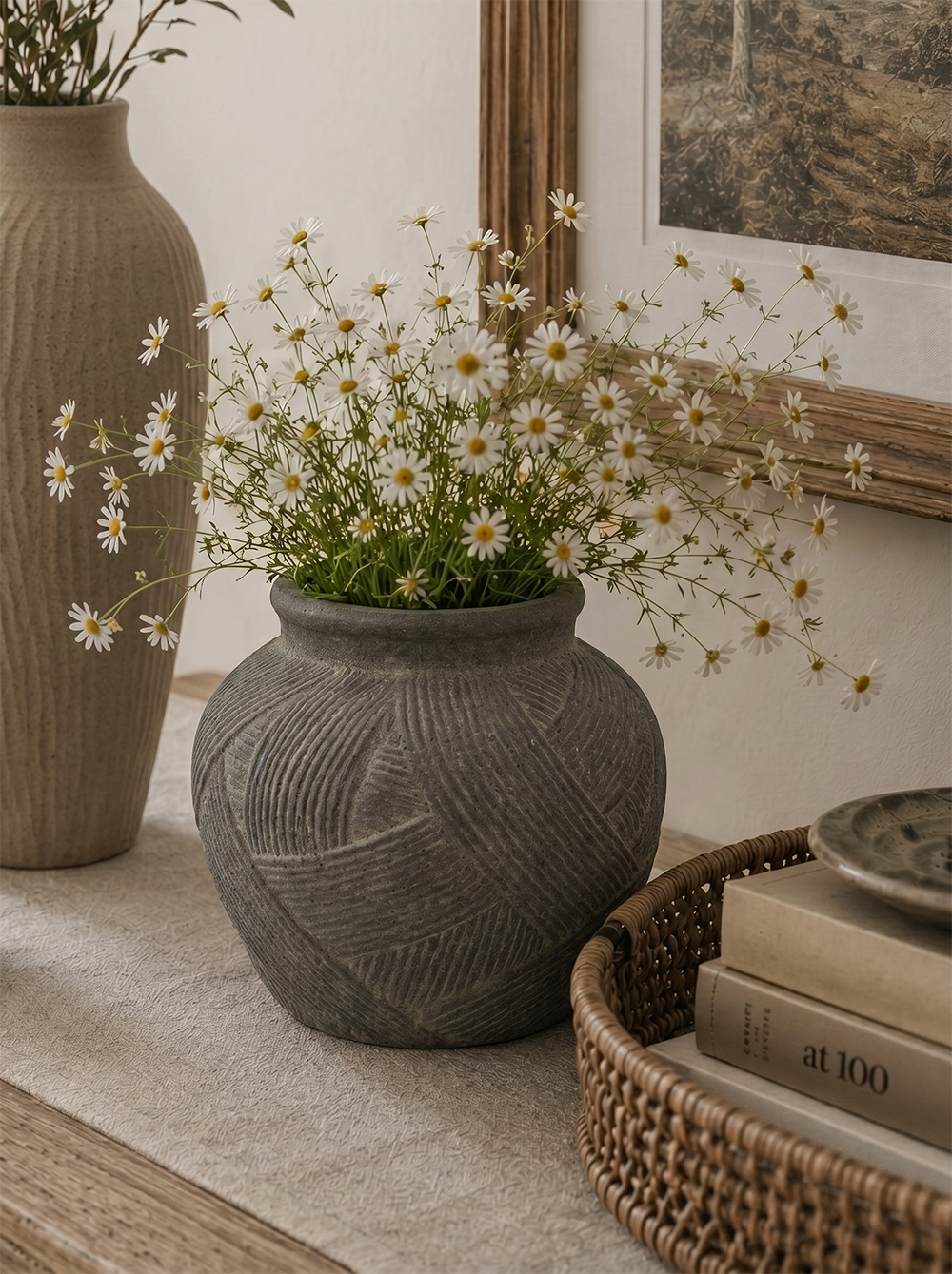 A gray textured ceramic pot filled with white daisies, styled on a beige table runner next to a woven basket and a tall beige vase.