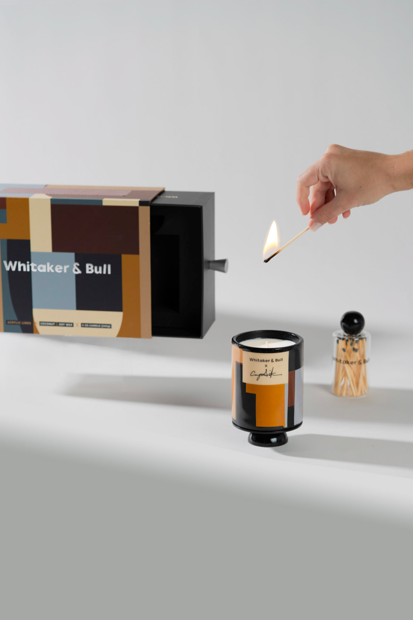 Candle being lit with a match, next to a decorative box and bottle on a light gray background