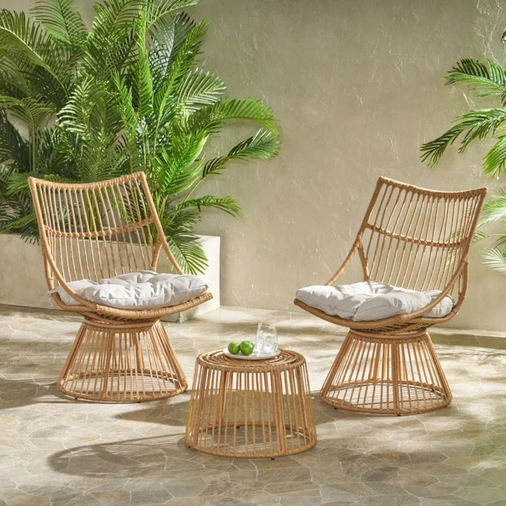 Two wicker chairs with white cushions and a matching table on a stone patio with plants in the background.
