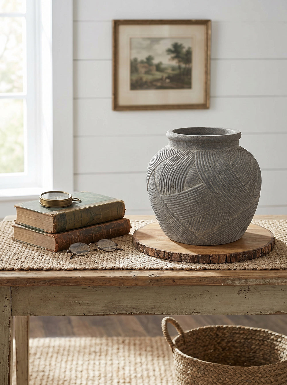 A gray textured ceramic vase with carved geometric patterns sits on a round wooden slice coaster on a rustic table. The table is styled with vintage books, a compass, and a woven placemat in a bright, modern room with white shiplap walls.