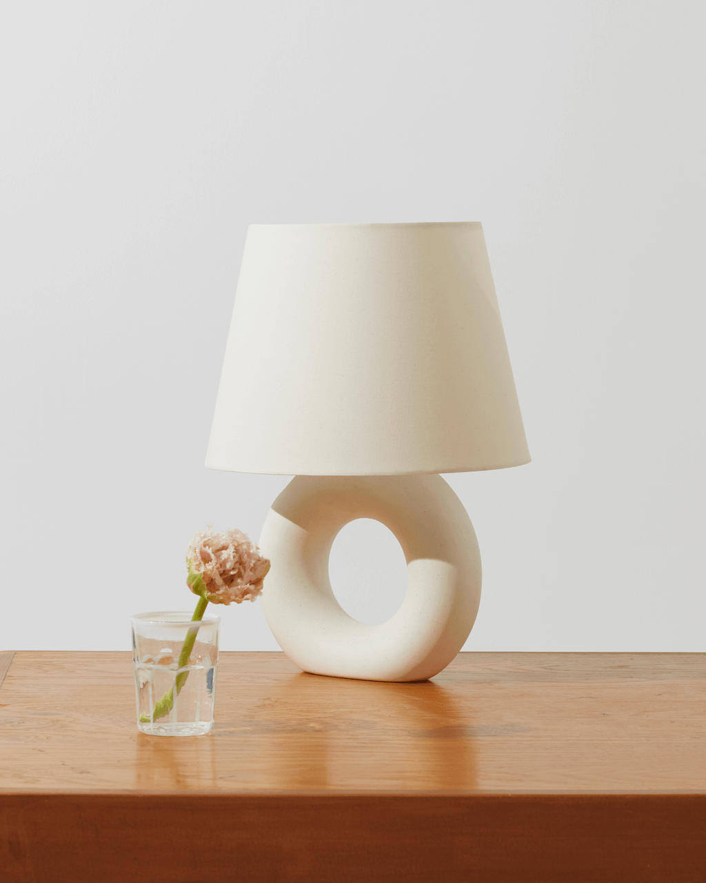 White table lamp with a unique donut base on a wooden surface, next to a glass with a flower.