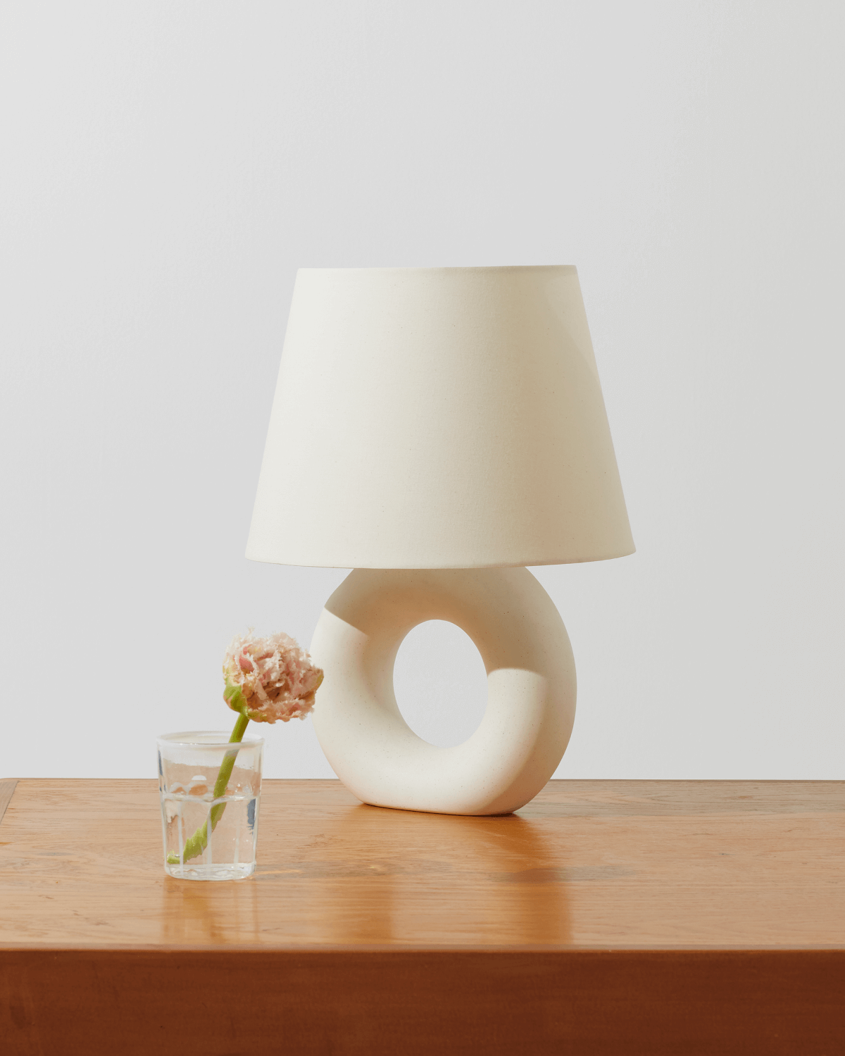 White table lamp with a unique donut base on a wooden surface, next to a glass with a flower.
