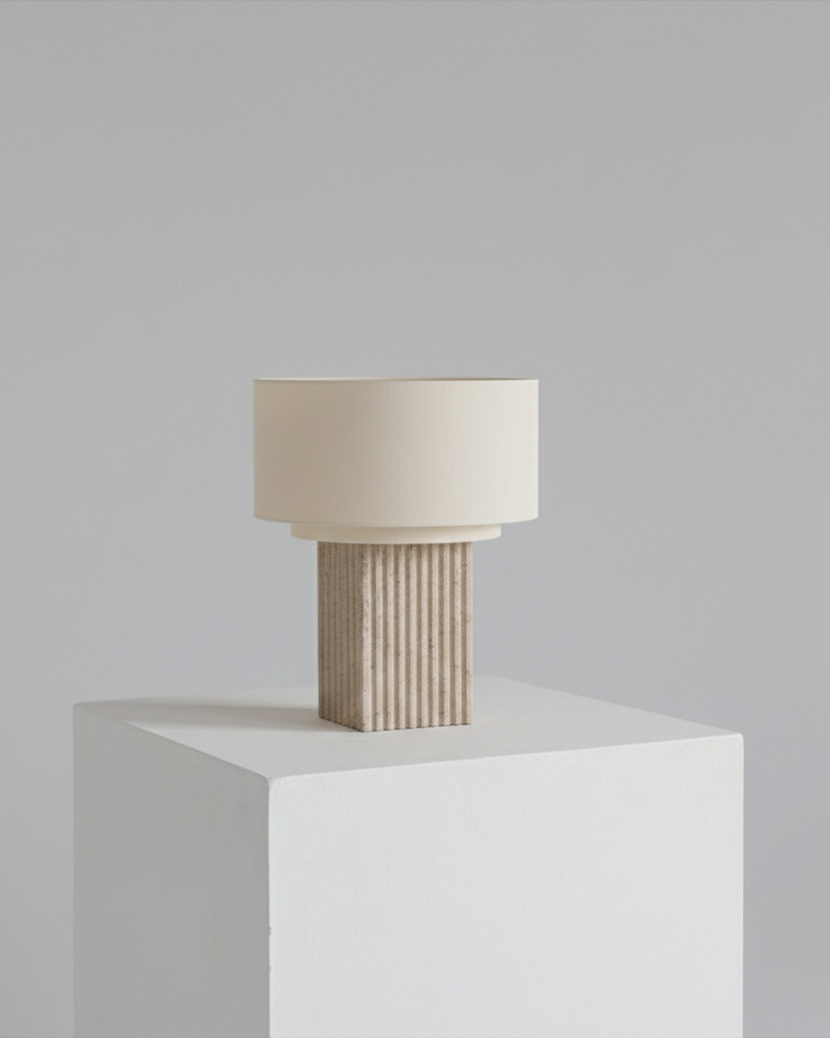Table lamp with a textured base and beige shade on a white pedestal against a light gray background