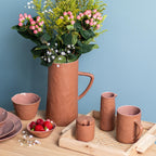 Set of terracotta ceramic items including a pitcher, cups, and a vase with flowers on a wooden surface against a blue wall.