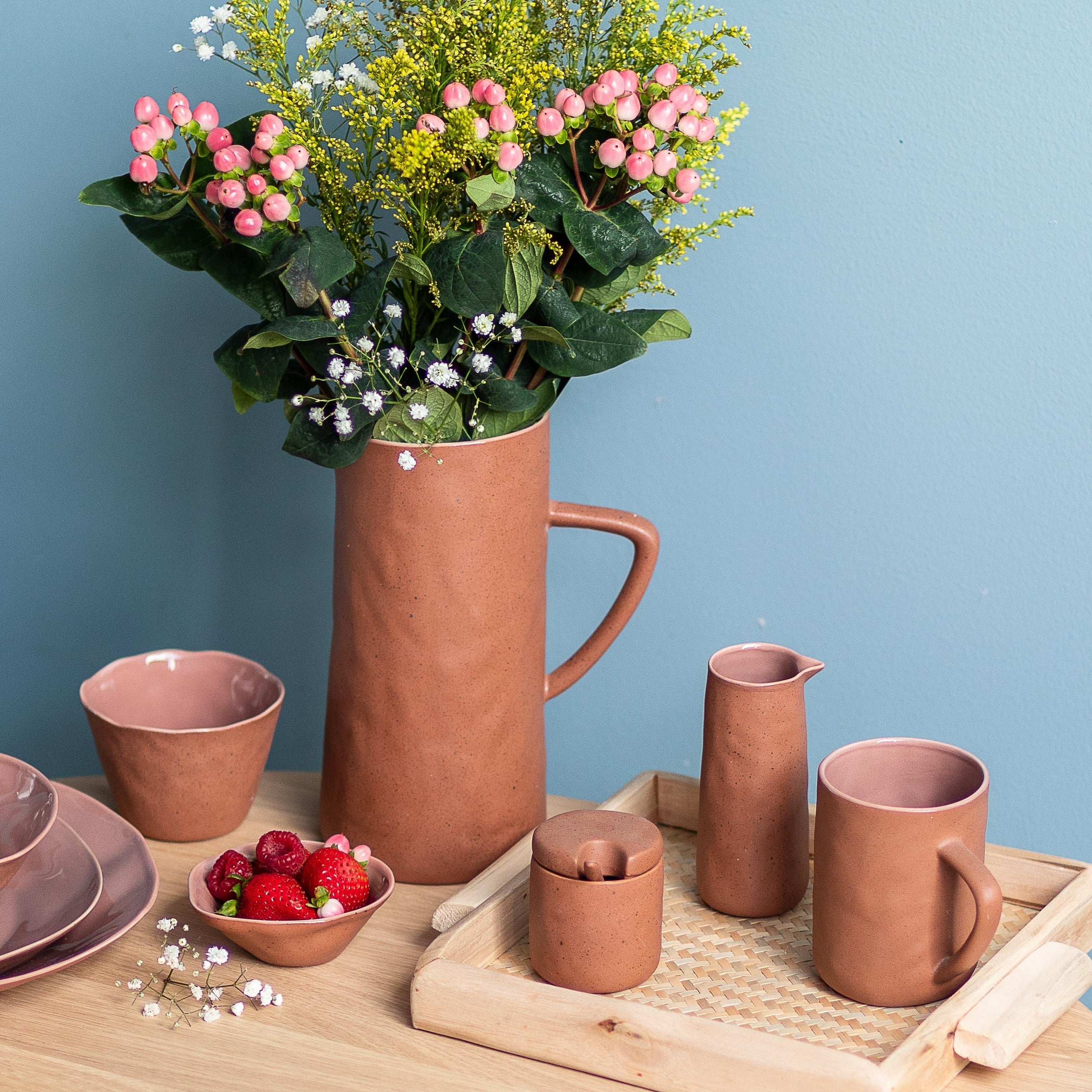 Set of terracotta ceramic items including a pitcher, cups, and a vase with flowers on a wooden surface against a blue wall.