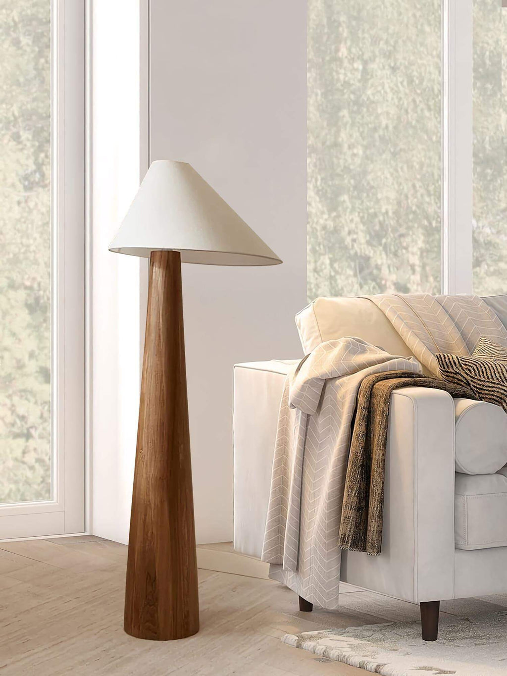 Floor lamp with wooden base and white shade next to a sofa with blankets in a room with large windows.