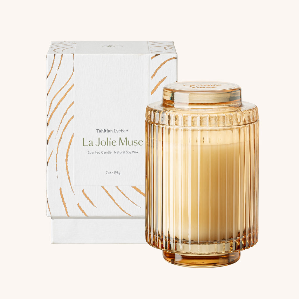 Glass candle with ribbed design and gold lid next to its packaging on a white background