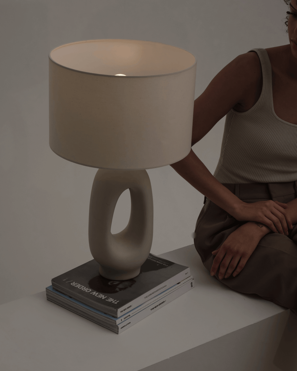 Person sitting next to a modern table lamp on a white surface with a neutral background