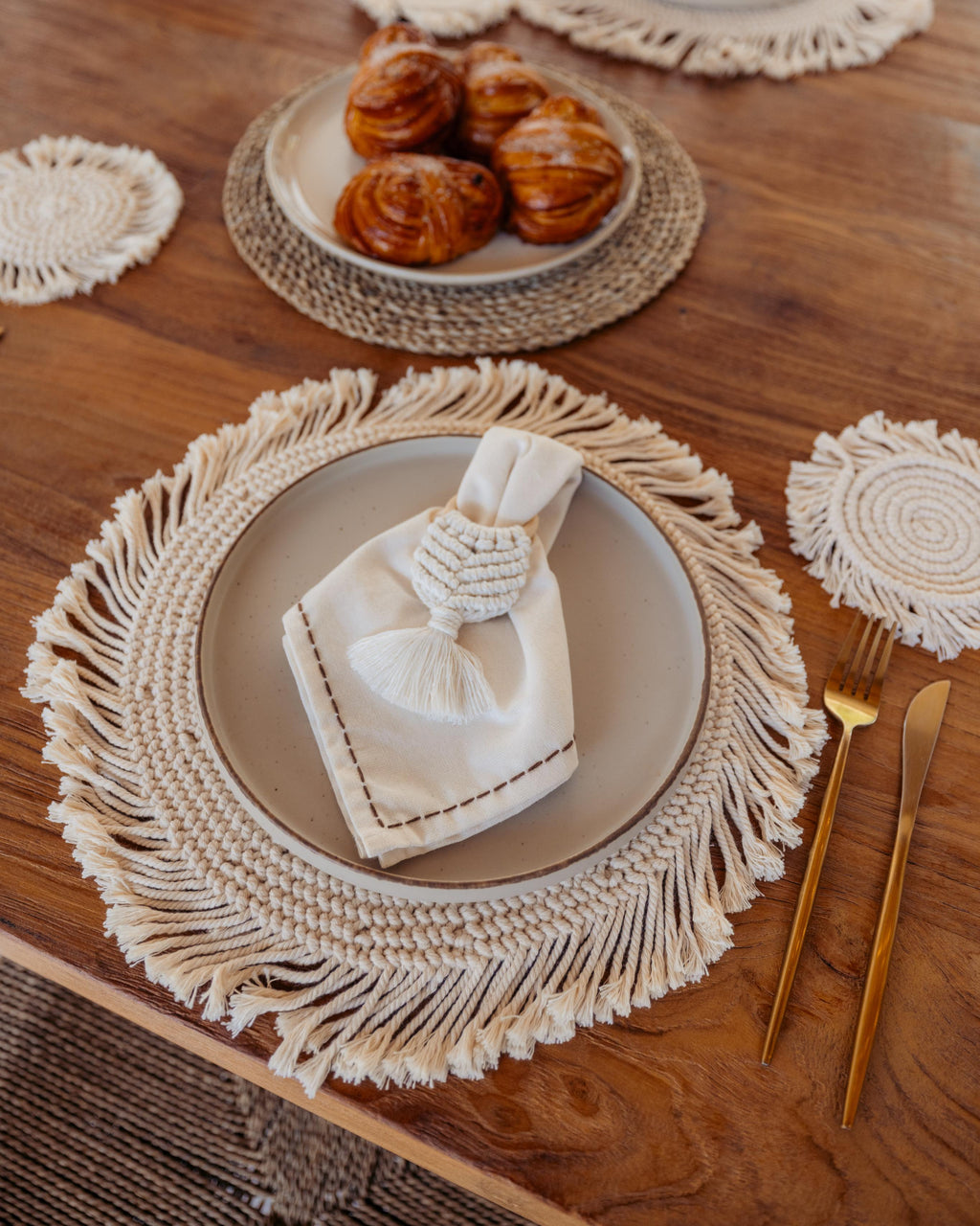 Table setting with macrame placemats, gold cutlery, and a plate of pastries.