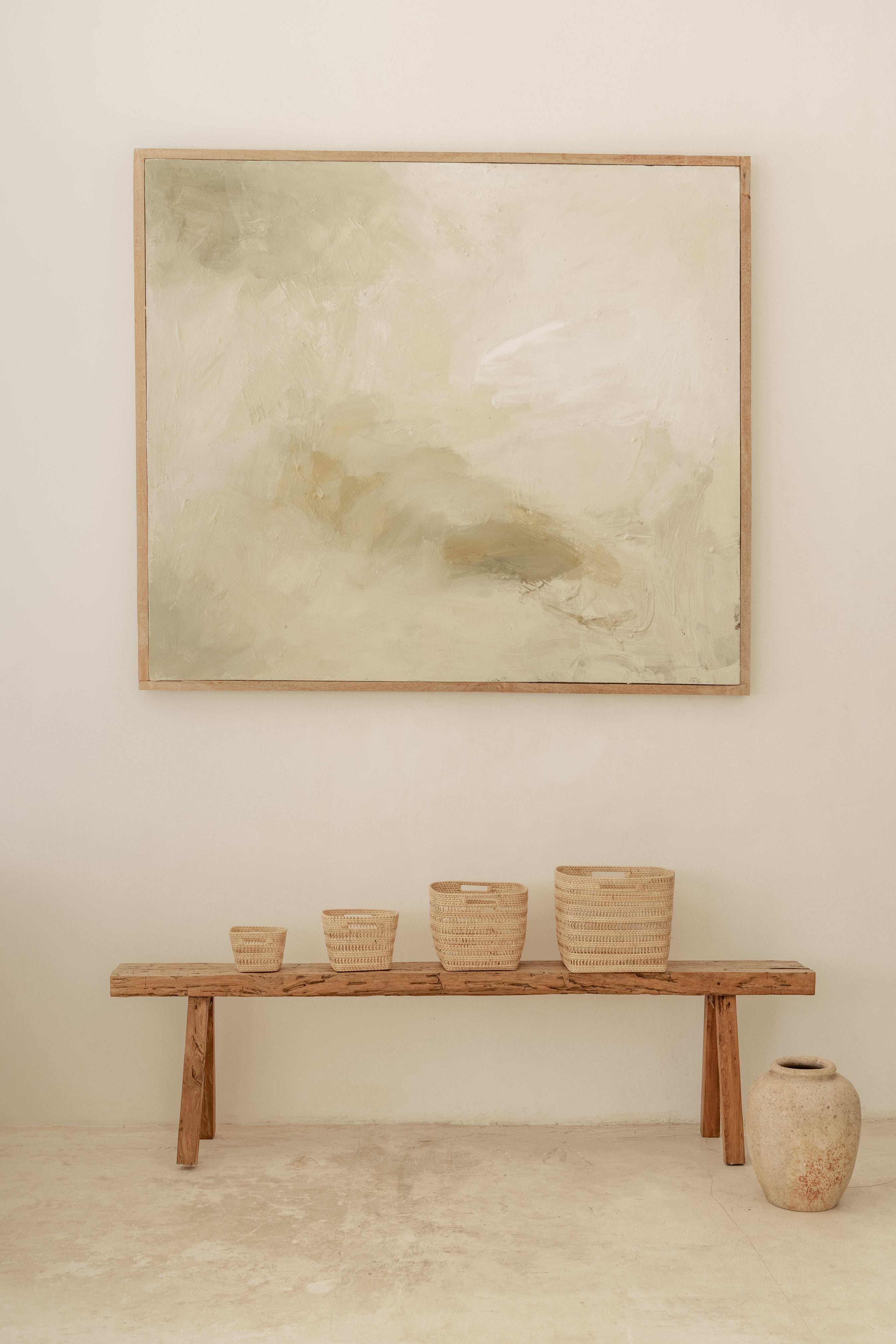 Wooden bench with woven baskets and a large abstract painting on a beige wall.