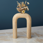 Wooden arch-shaped vase with dried flowers on a marble surface against a dark blue wall.