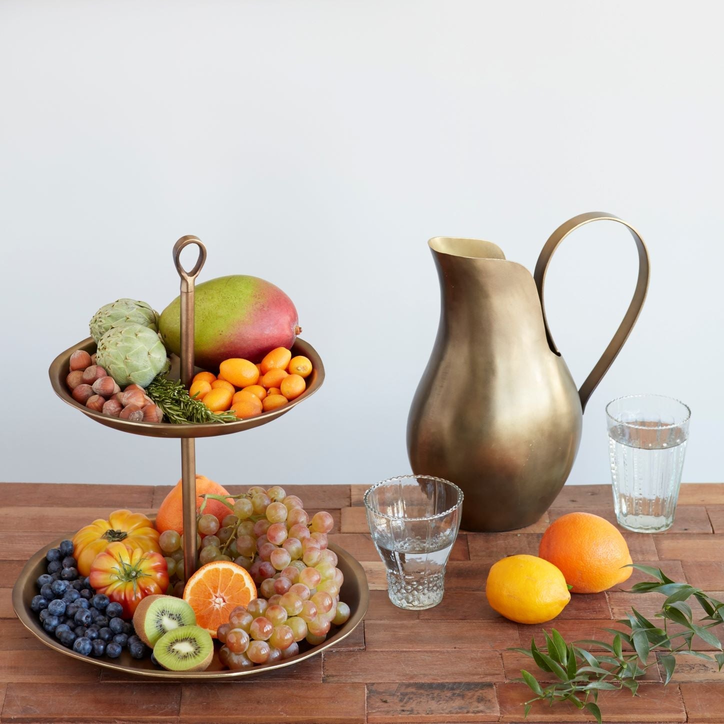 Fruit stand with fruits and a brass pitcher on a wooden table.