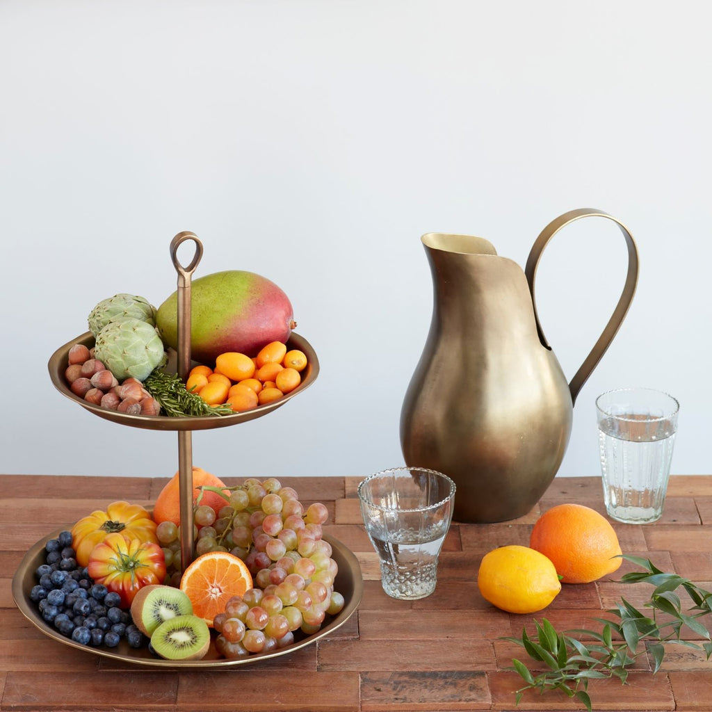 Fruit stand with fruits and a brass pitcher on a wooden table.