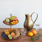 Fruit stand with fruits and a brass pitcher on a wooden table.