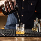 Barista pouring drink into a glass on a bar counter with various barware tools.