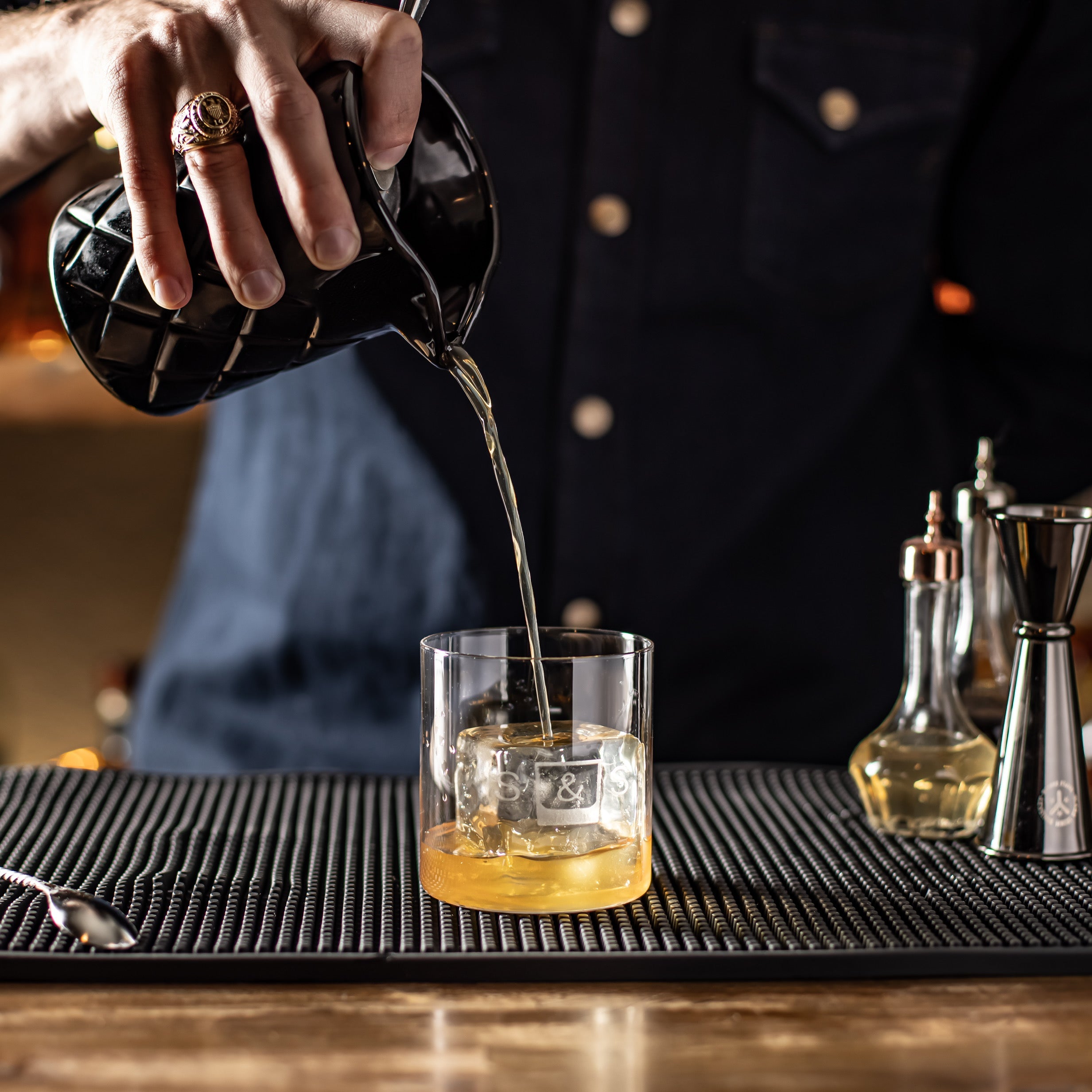 Barista pouring drink into a glass on a bar counter with various barware tools.