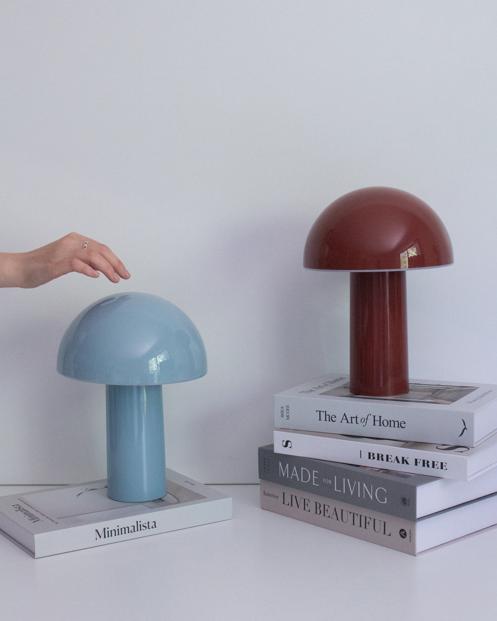 Two mushroom-shaped lamps, one blue and one brown, on a stack of books with a hand reaching towards the blue lamp.