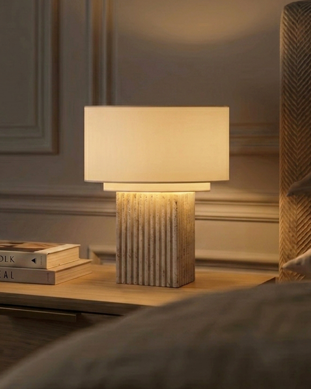 A table lamp with a rectangular cream fabric shade and a ribbed, textured light wood base, illuminated on a wooden bedside table in a warm, elegant room setting.