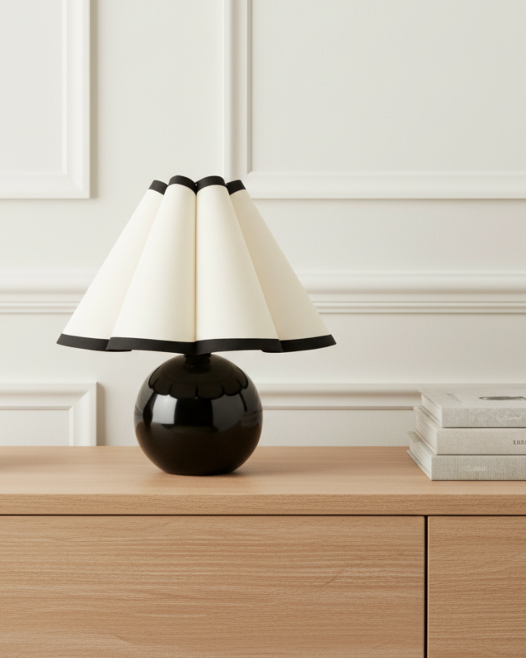 A modern table lamp with a glossy black spherical base and a pleated cream shade, styled on a light wood console table against a white paneled wall.