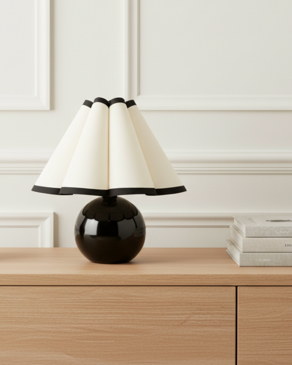 A modern table lamp with a glossy black spherical base and a pleated cream shade, styled on a light wood console table against a white paneled wall.