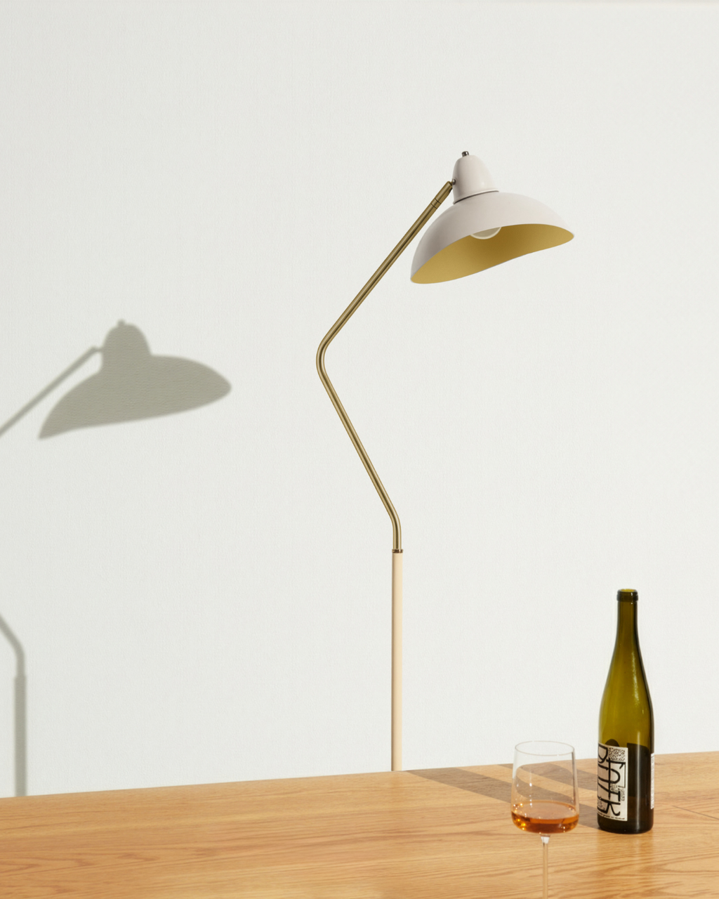 A modern floor lamp with a curved brass arm and a white dome shade with a gold interior, standing on a light wood table next to a wine bottle and a glass of red wine against a white wall.