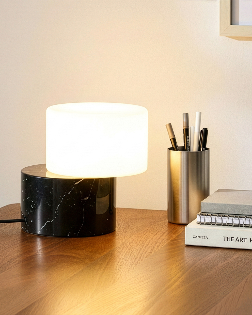 A modern table lamp with a glowing white cylindrical shade and a glossy black marble base, styled on a wooden desk next to a metal pen cup and books.
