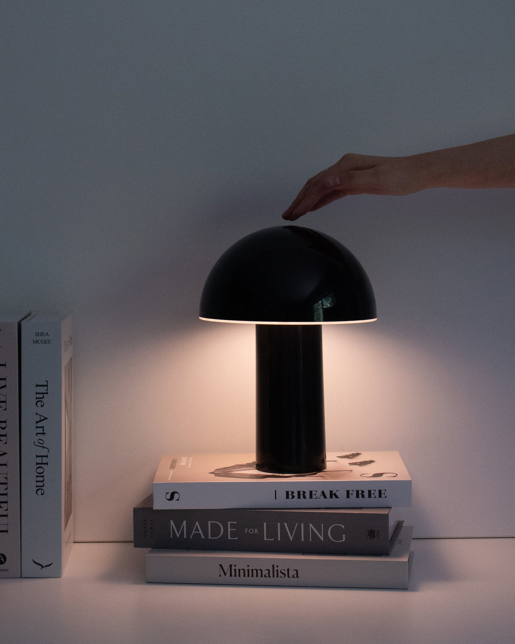 Black table lamp being turned on with a hand, placed on a stack of books against a light background.