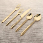 Set of gold-colored cutlery including a knife, two forks, and two spoons on a textured beige surface.