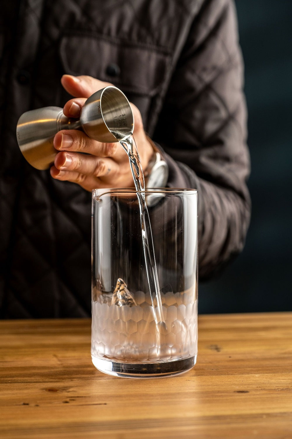 Person pouring liquid from a jigger into a glass on a wooden surface