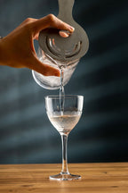 Hand pouring liquid from a clear container into a wine cocktail glass on a wooden surface with a dark background