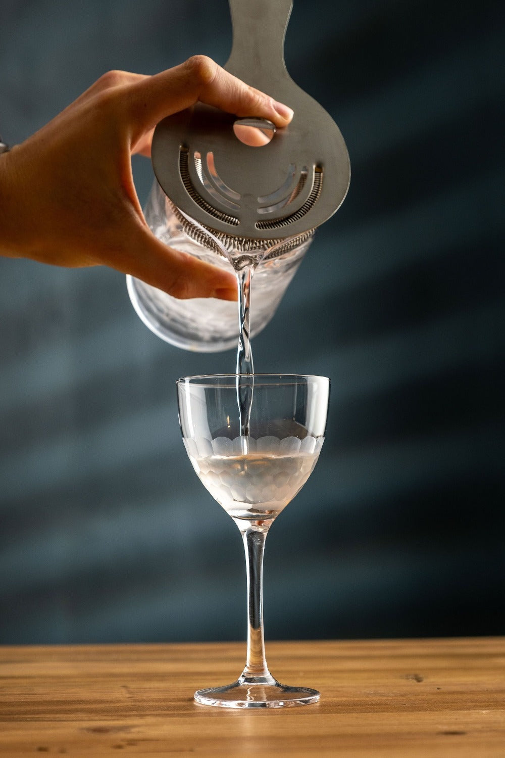 Hand pouring liquid from a clear container into a wine cocktail glass on a wooden surface with a dark background