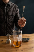 Person stirring a cocktail in a glass with ice on a wooden table.