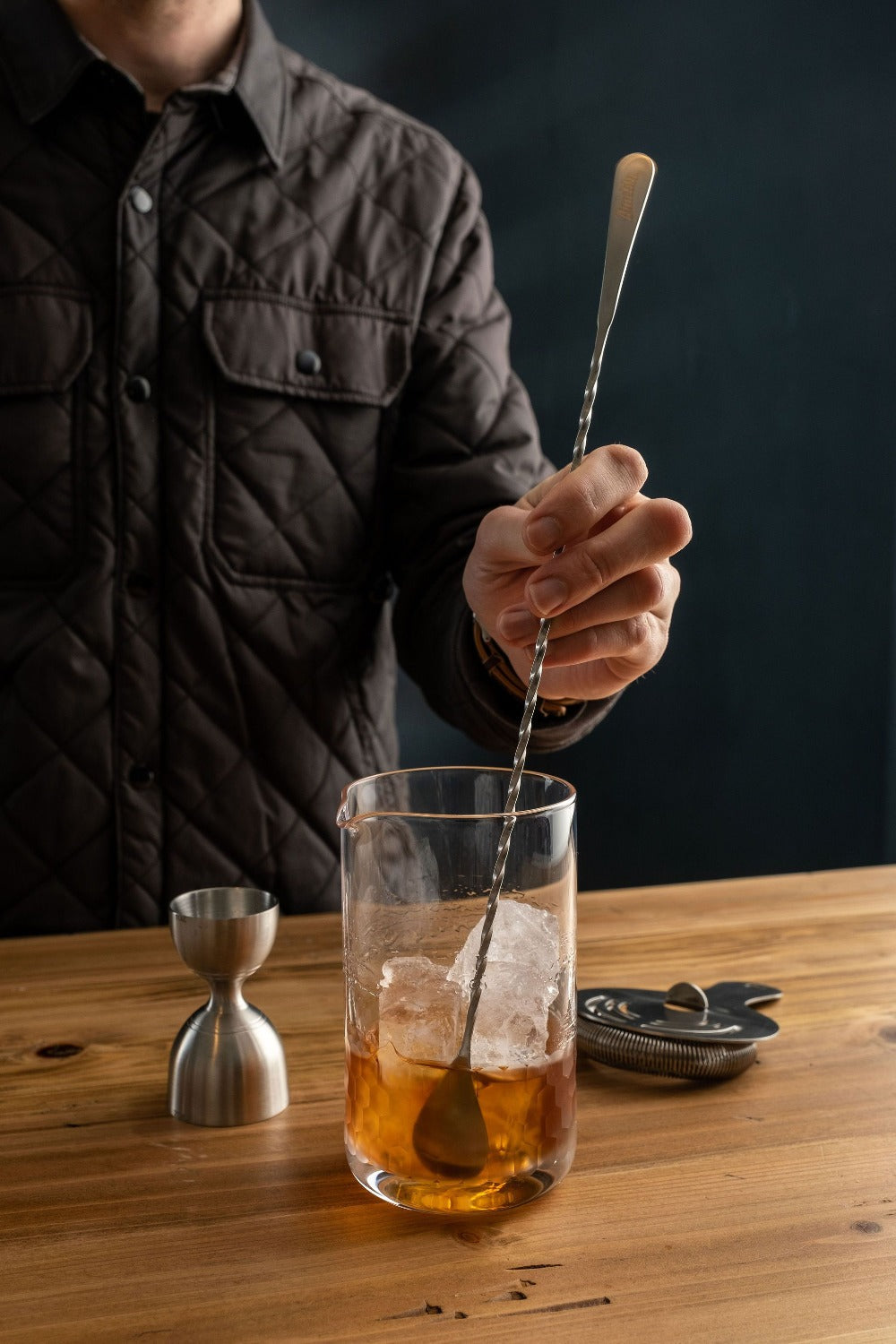 Person stirring a cocktail in a glass with ice on a wooden table.
