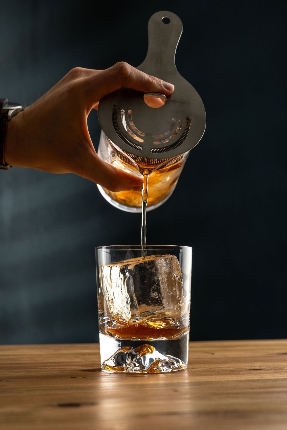 Hand pouring liquid from a glass container into a glass with ice on a wooden surface.
